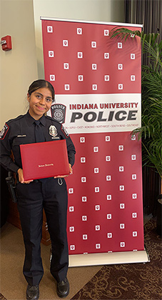 Ingrid Ortega in uniform holding her diploma in ballroom in front of vertical Indiana University Police banner